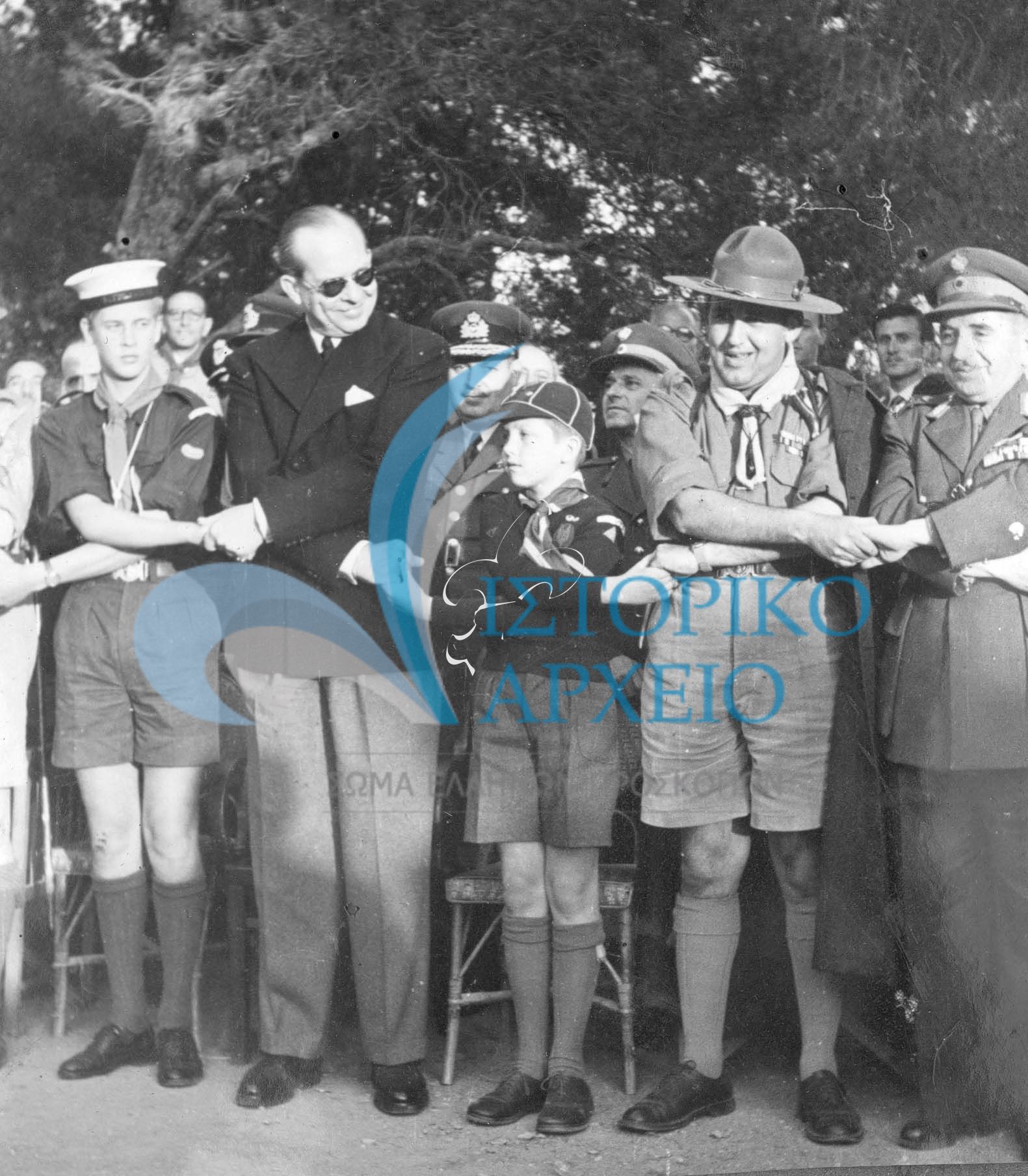 Εορταστική πυρά για την Δ` Συνάντηση Πτυχιούχων Διακριτικού Δάσους στο Προσκοπικό Κέντρο Πύργου Βασιλίσσης το 1951. Από αριστερά: Πρίγκιπας Κάρολος, Βασιλιάς Παύλος, Διάδοχος Κωνσταντίνος με στολή λυκόπουλου, και ο Δ. Αλεξάτος. 