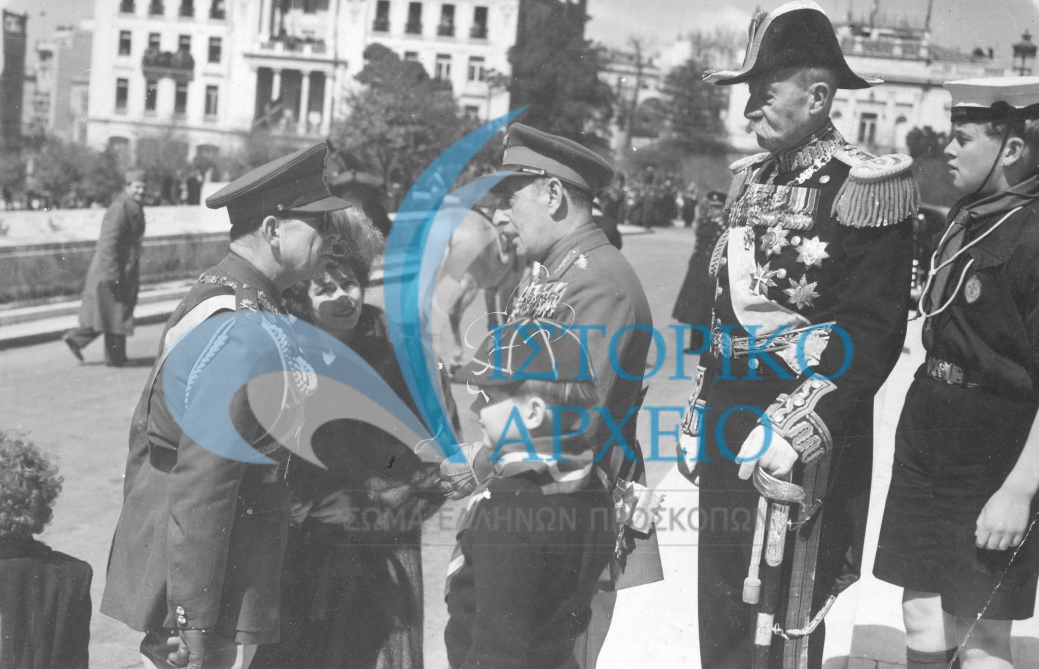 Ο Βασιλιάς Παύλος χαιρετάει τον Στρατάρχη Παπάγου λίγο πριν τη παρέλαση της 25ης Μαρτίου του 1949. Δίπλα του ο Διάδοχος Κωνσταντίνος με στολή λυκόπουλου. Πλησίον παρακολουθεί ο πρίγκιππας Ανδρέας με τον εγγονό του ναυτοπρόσκοπο Αριγένη Κάρολο.