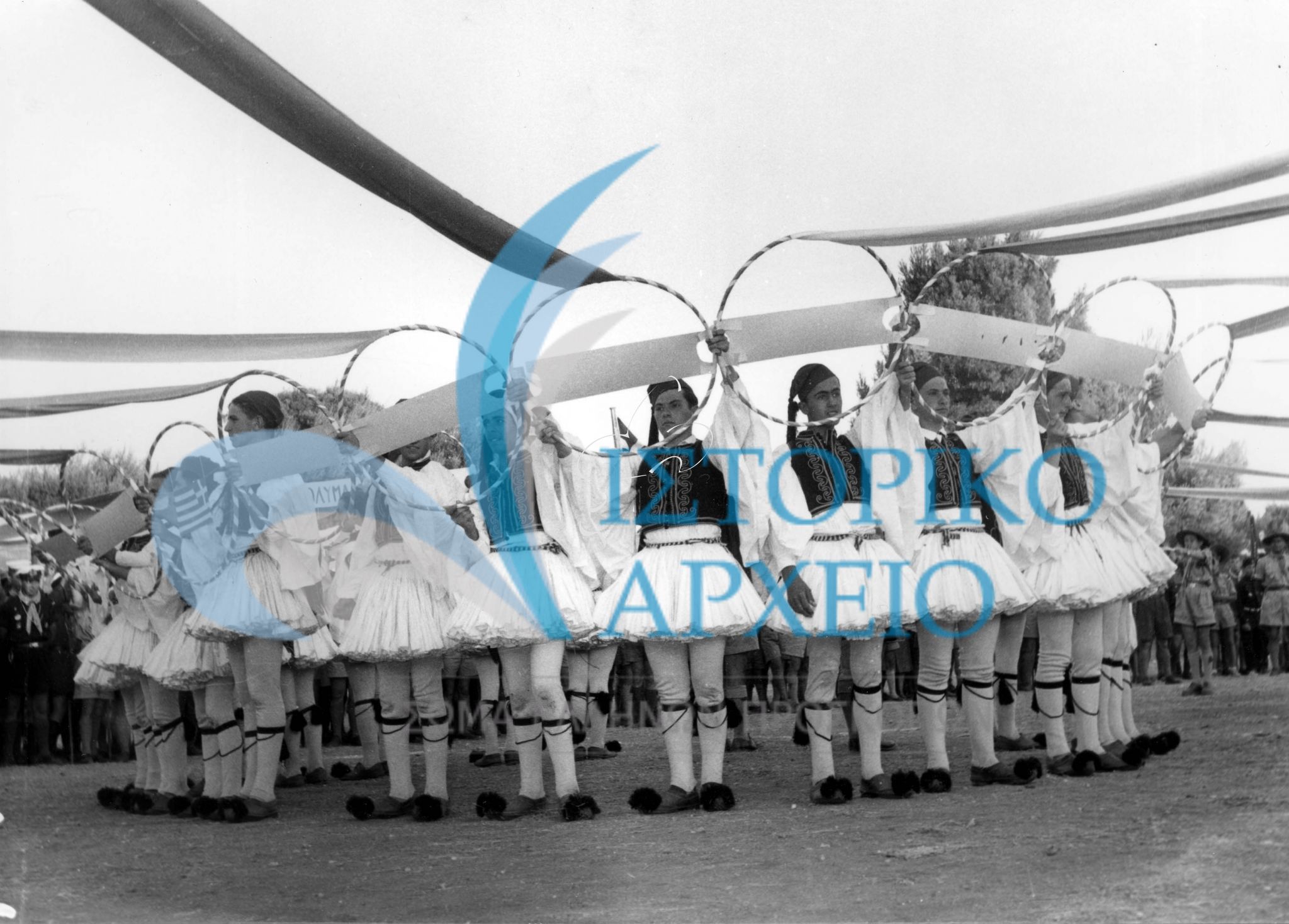 Έλληνες πρόσκοποι από όλο το κόσμο φορώντας εθνική ενδυμασία συνδέουν τους κύκλους των πόλεων όπου βρίσκονται ελληνικά προσκοπικά συστήματα κατά την Πυρά Έναρξης του Πανελληνίου Τζάμπορη Αποδήμων τον Αύγουστο του 1956.