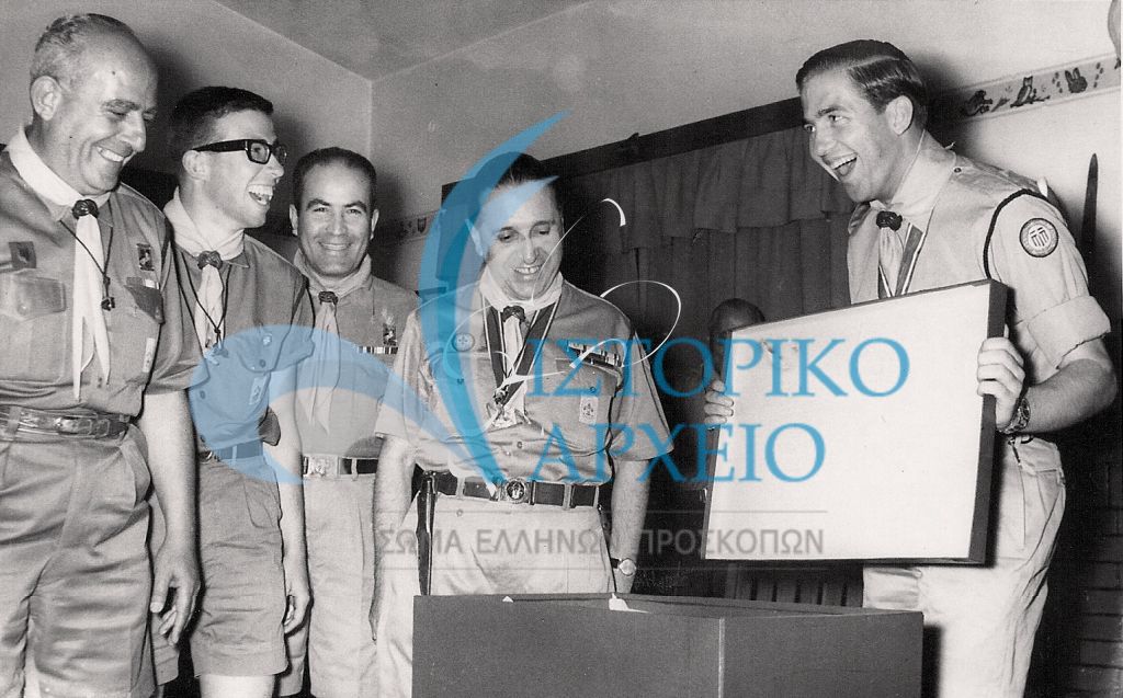 Ο Διάδοχος Κωνσταντίνος ανοίγει ενθουσιασμένος το δώρο του μετά τη Συνάντηση των Πτυχιούχων Διακριτικού Δάσους το 1963. Στο επιτελείο ο έφορος εκπαιδεύσεως Δ. Μακρίδης.