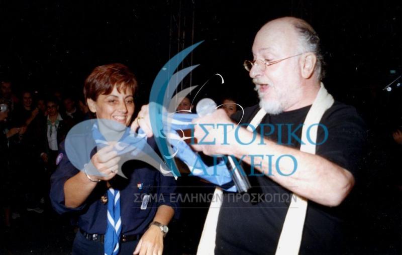 Η έφορος Δημοσίων Σχέσεων Δ. Παπαθεωδούλου ξαναφοράει το προσκοπικό μαντήλι στον Διονύση Σαββόπουλο σε συναυλία για τον εθελοντισμό των Ολυμπιακών Αγώνων "Αθήνα 2004" στην Πάτρα.