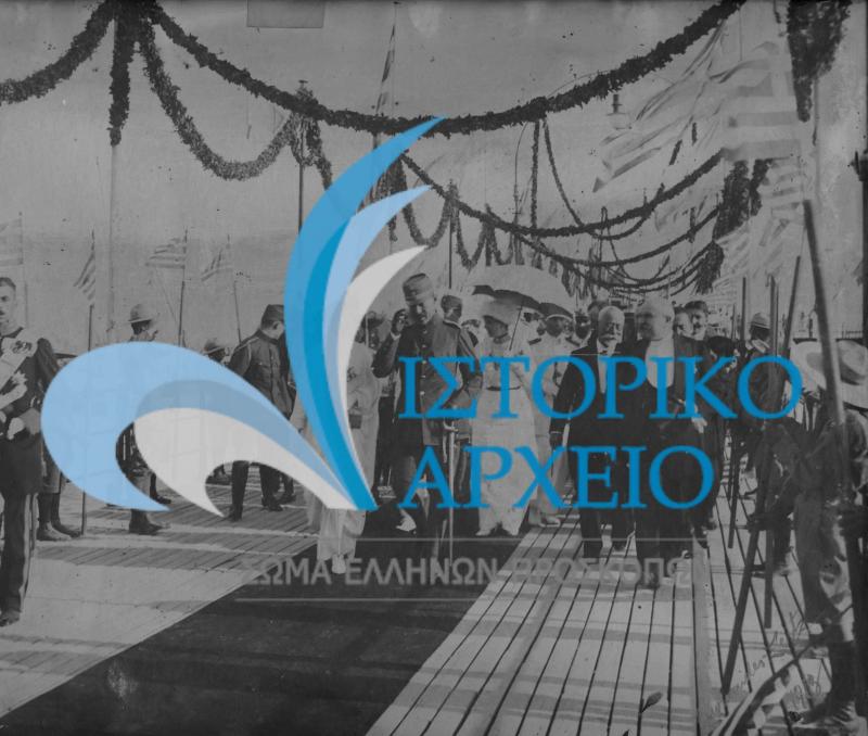 Οι πρώτοι Έλληνες Πρόσκοποι ως τιμητικό άγημα με υψωμένα κοντάρια στην επίσημη υποδοχή του Βασιλιά Κωνσταντίνου στο Νέο Φάληρο το 1913 μετά τους Βαλκανικούς Πολέμους. Διακρίνονται επίσης: η Βασίλισσα Σοφία, η Πριγκίπισσα Ελένη, η Βασίλισσα Ειρήνη, ο Πρίγκιπας Παύλος, ο Υπουργός Στρατιωτικών Στράτος, ο Πρωθυπουργός Ελ. Βενιζέλος, ο Υπουργός Εξωτερικών Μιχαλακόπουλος και ο Υπουργός Οικονομικών Κορομηλάς.