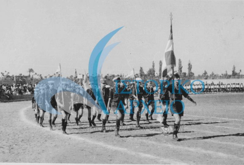 Οι Έλληνες Πρόσκοποι του Λιβάνου σε σε παρέλαση σε στάδιο στην Τρίπολη της Λιβύης κατά την διάρκεια προσκοπικών αγώνων το 1960. Σημαιοφόρος ο Π. Τσακμάκης με υπαρχηγό τον Λ. Κάη.